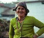 A joyful elderly woman wearing a green knitted sweater and colorful beanie smiles warmly outdoors.